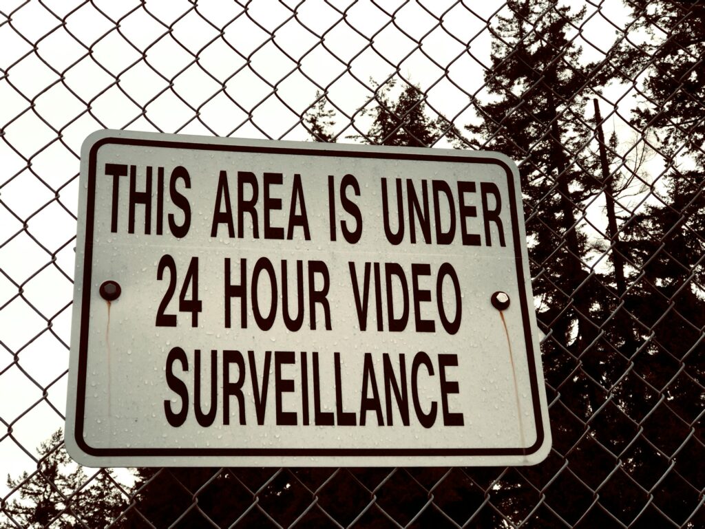 Warning sign reading, "THIS AREA IS UNDER 24 HOUR VIDEO SURVEILLANCE" on a chain link fence.