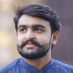 A thoughtful bearded man in blue attire poses outdoors in Kasur, highlighting cultural fashion.