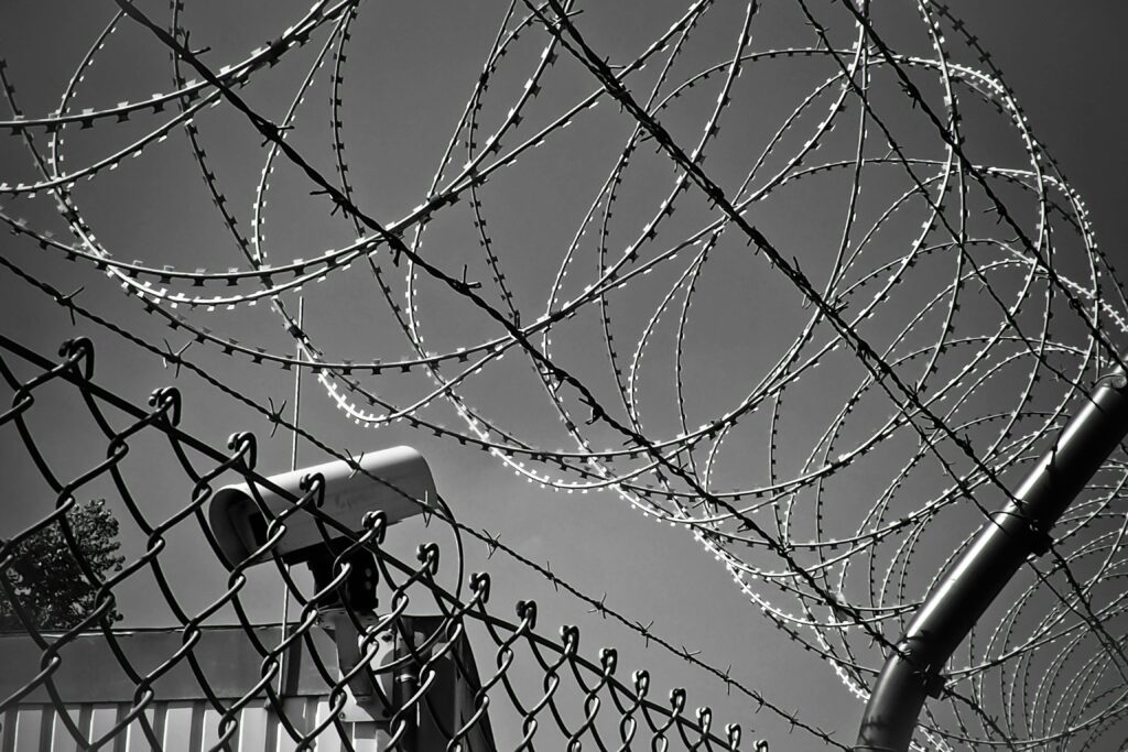 Security camera positioned behind a barbed wire fence in black and white, indicating high security.